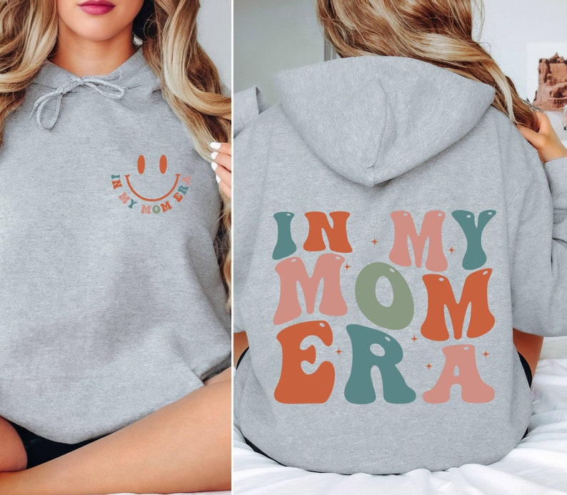 May include: Gray hooded sweatshirt with the text "In My Mom Era" in colorful retro font on the back. The front of the sweatshirt has a smiley face with the text "In My Mom Era" around it.