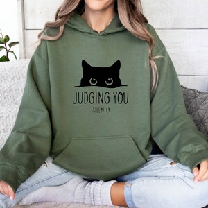 K&ouml;nnte beinhalten: Ein gr&uuml;ner Kapuzenpullover mit einer schwarzen Katze, die &uuml;ber den Rand schaut, und dem Text "JUDGING YOU SILENTLY".