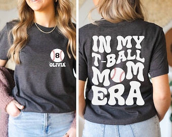 May include: A dark gray t-shirt with the text "In My T-Ball Mom Era" in white, retro-style lettering. A white baseball with red stitching is centered above the text. The shirt has a pocket on the front with a white baseball and the number "8" and the name "Olivia" in black.