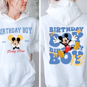 K&ouml;nnte beinhalten: Wei&szlig;er Hoodie mit einem blauen und gelben Motiv von Mickey Mouse auf der R&uuml;ckseite. Der Text "Birthday Boy" ist in blau und gelb auf der R&uuml;ckseite. Die Vorderseite des Hoodies hat ein kleineres Motiv von Mickey Mouse mit dem Text "Birthday Boy" in blau und gelb, "Est 2011" in schwarz und "Mickey Mouse" in rot.