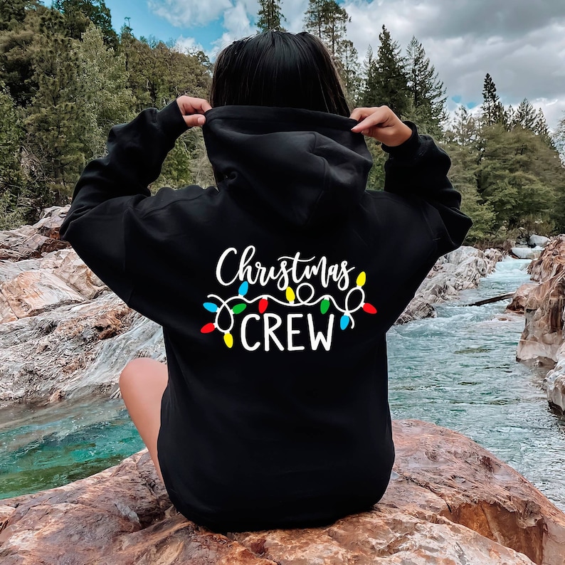 May include: Black hooded sweatshirt with white text that reads "Christmas Crew" and a string of colourful Christmas lights.