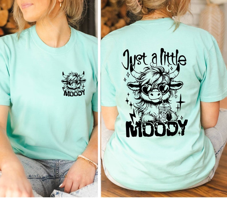 Puede incluir: Una camiseta turquesa con un gr&aacute;fico negro de una vaca con gafas de sol y sosteniendo una bebida. El texto "Just a little MOODY" est&aacute; impreso en la parte posterior de la camiseta.