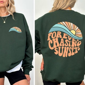 May include: Dark green sweatshirt with a small sunset graphic on the front and a larger "Forever Chasing Sunsets" design on the back. The design features a sun, waves, and text in orange, blue, and yellow. The sweatshirt is a casual style.