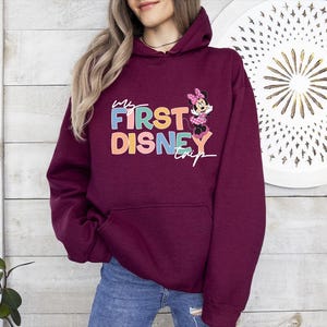 May include: Burgundy hooded sweatshirt with the text "My First Disney Trip" in colorful letters and a Minnie Mouse graphic. The sweatshirt has a front pocket and a drawstring hood.