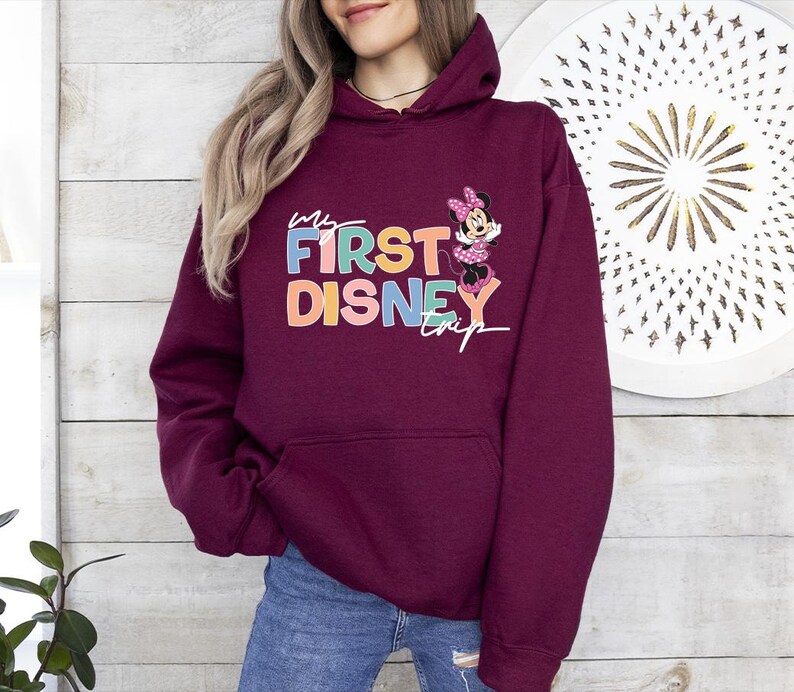May include: Burgundy hooded sweatshirt with the text "My First Disney Trip" in colorful letters and a Minnie Mouse graphic. The sweatshirt has a front pocket and a drawstring hood.