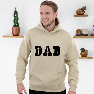 Puede incluir: Una sudadera con capucha beige con la palabra "DAD" impresa en negro en la parte delantera. Las letras est&aacute;n estilizadas con un martillo y una llave inglesa.