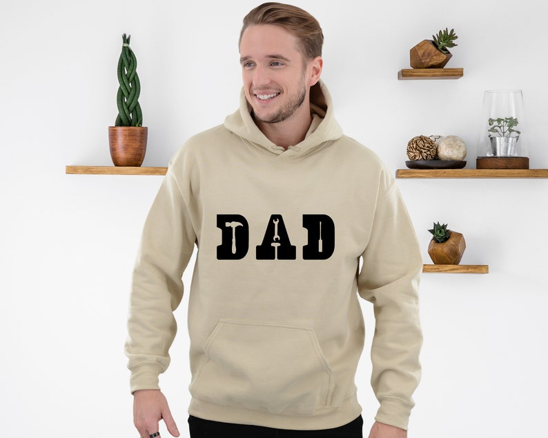 Puede incluir: Una sudadera con capucha beige con la palabra "DAD" impresa en negro en la parte delantera. Las letras est&aacute;n estilizadas con un martillo y una llave inglesa.