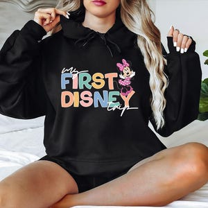 May include: Black hooded sweatshirt with the text "My First Disney Trip" in colorful letters. A Minnie Mouse graphic is on the right side. The sweatshirt is a comfortable, casual style.