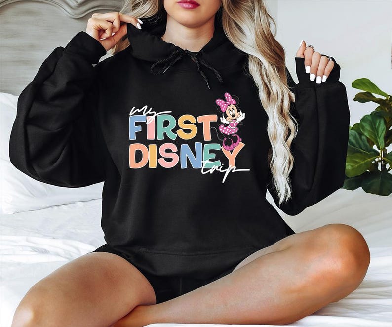 May include: Black hooded sweatshirt with the text "My First Disney Trip" in colorful letters. A Minnie Mouse graphic is on the right side. The sweatshirt is a comfortable, casual style.