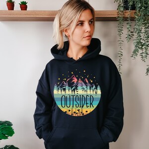 May include: A navy blue hooded sweatshirt featuring a colorful mountain and forest design within a circular shape. The word "OUTSIDER" is printed in white. The design includes stars, trees, and a striped background.