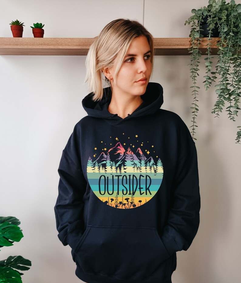 May include: A navy blue hooded sweatshirt featuring a colorful mountain and forest design within a circular shape. The word "OUTSIDER" is printed in white. The design includes stars, trees, and a striped background.