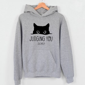 K&ouml;nnte beinhalten: Ein grauer Kapuzenpullover mit einer schwarzen Katze, die &uuml;ber den Rand schaut, und dem Text "JUDGING YOU SILENTLY."