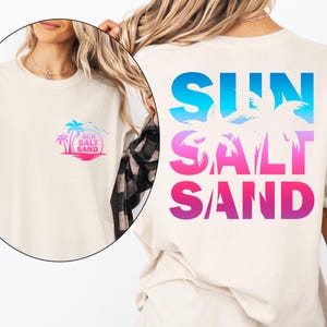 Pode incluir: Camiseta creme com design tropical. A frente apresenta um pequeno gr&aacute;fico de uma palmeira, sol e as palavras "Sun Salt Sand". A parte de tr&aacute;s exibe as mesmas palavras em letras grandes gradientes azuis, rosas e roxas com silhuetas de palmeiras.