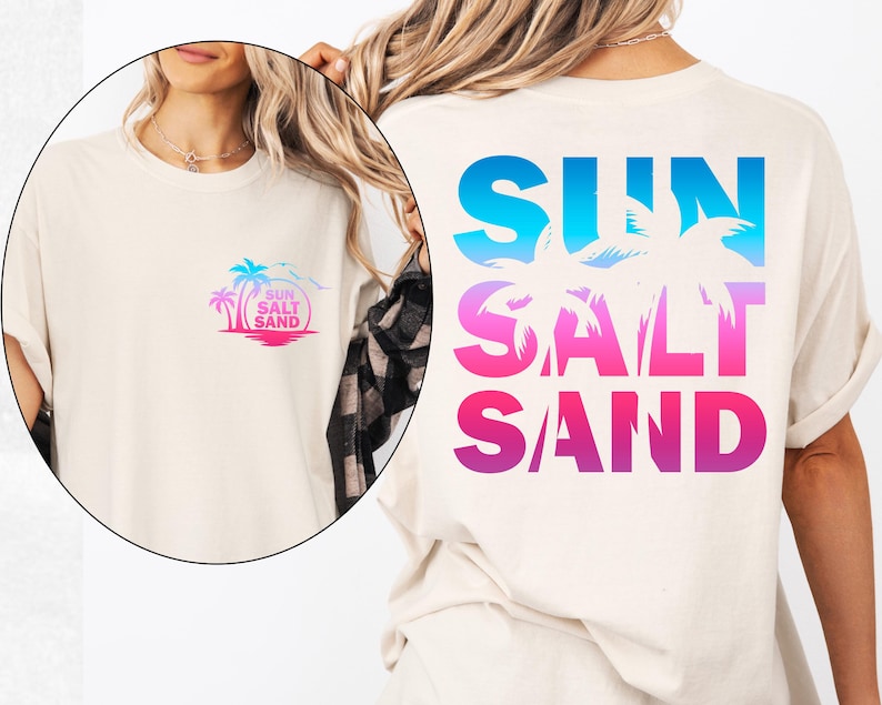 Pode incluir: Camiseta creme com design tropical. A frente apresenta um pequeno gr&aacute;fico de uma palmeira, sol e as palavras "Sun Salt Sand". A parte de tr&aacute;s exibe as mesmas palavras em letras grandes gradientes azuis, rosas e roxas com silhuetas de palmeiras.