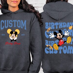 K&ouml;nnte beinhalten: Ein dunkelgrauer Hoodie mit einem blauen und wei&szlig;en Mickey-Mouse-Motiv auf der Vorderseite. Das Motiv lautet "Custom Est 2011 Mickey Mouse". Die R&uuml;ckseite des Hoodies hat ein blaues und wei&szlig;es Motiv mit der Aufschrift "Birthday Custom Birthday Custom".