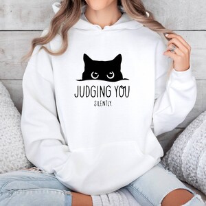 K&ouml;nnte beinhalten: Wei&szlig;er Kapuzenpullover mit einer schwarzen Katze, die &uuml;ber den Rand schaut, und dem Text "JUDGING YOU SILENTLY."