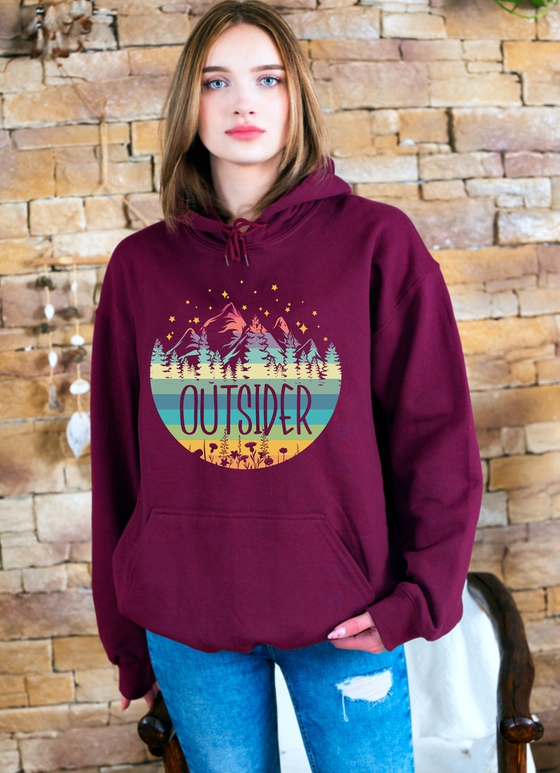 May include: Burgundy hoodie with a front pocket and a drawstring hood. The graphic on the front features a mountain scene with the word "OUTSIDER" in a circular design. The design includes mountains, trees, and stars.