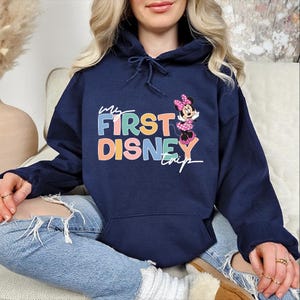 May include: Navy blue hooded sweatshirt with the text "My First Disney Trip" in colorful block letters. A Minnie Mouse graphic is on the right side. The sweatshirt has a drawstring hood and a front pocket.