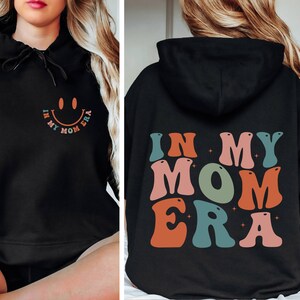May include: Black hooded sweatshirt with the text "In My Mom Era" in colorful retro letters. A smiley face is on the front of the sweatshirt.