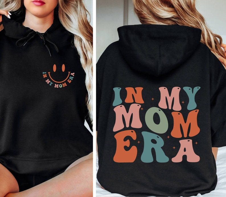 May include: Black hooded sweatshirt with the text "In My Mom Era" in colorful retro letters. A smiley face is on the front of the sweatshirt.