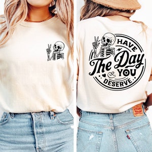May include: A white t-shirt with a black skeleton graphic on the front and a black circle graphic on the back that says "Have the Day You Deserve".