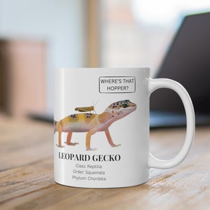 Puede incluir: Taza de cerámica blanca con un gecko leopardo de dibujos animados y un saltamontes en su espalda. La taza tiene el texto "Where's that hopper?" y "Leopard Gecko" con su clasificación científica.