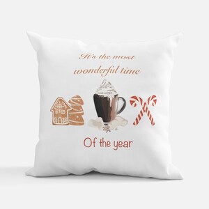 Può includere: Cuscino decorativo bianco con un design a tema festivo. Il design include una casa e un albero di pan di zenzero, una tazza di cioccolata calda con panna montata, marshmallow, bastoncini di zucchero e il testo "It's the most wonderful time of the year."