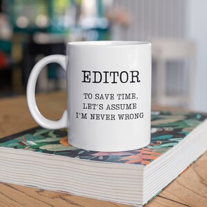 May include: White ceramic mug with the word "EDITOR" in black capital letters. Below the word, the text reads: "TO SAVE TIME, LET'S ASSUME I'M NEVER WRONG." The mug is on top of a book with a floral design.
