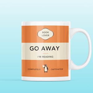 Puede incluir: Taza de cerámica blanca con un diseño naranja. La taza presenta el texto "Book Lover", "Go Away I'm Reading", "Completely", y "Captivated". Una ilustración de pingüino se encuentra entre las palabras "Completely" y "Captivated".