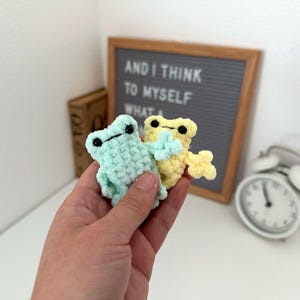 May include: Two small, handmade crochet frogs in pastel colors. One is light blue, the other is yellow. Both have black eyes and are held in a hand. The frogs are a whimsical craft item.