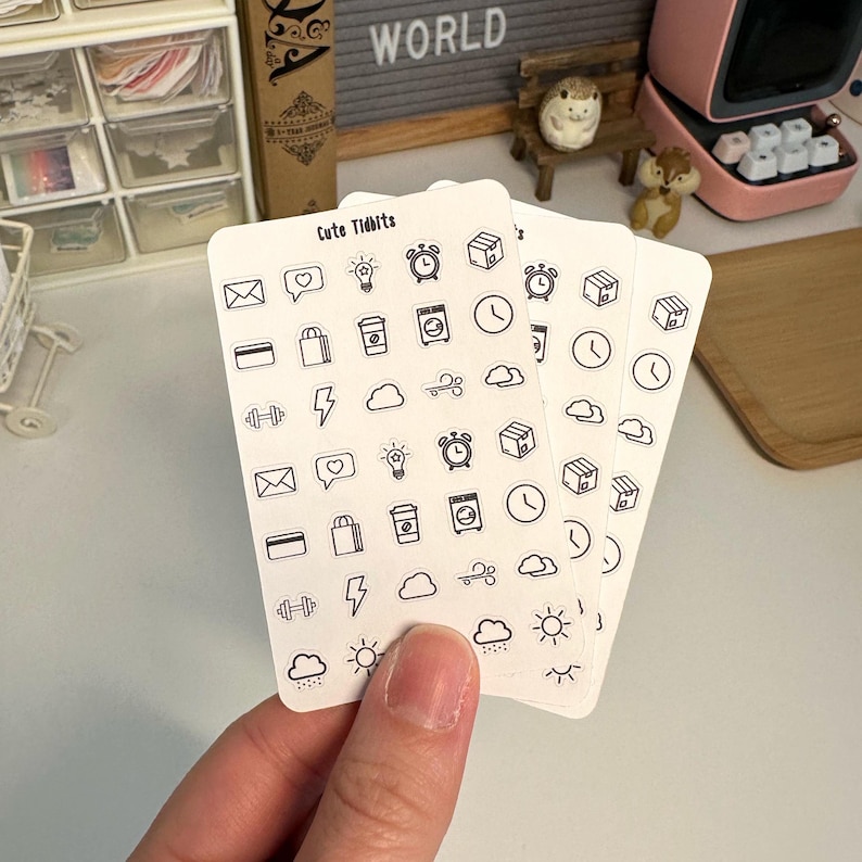 May include: A set of four sheets of clear stickers with black outlines of various icons, including a lightbulb, a coffee cup, a cloud, a lightning bolt, a shopping bag, a dumbbell, a clock, a package, a heart, an envelope, a sun, and a rain cloud. The stickers are labeled "Cute Tidbits".