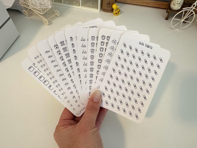 May include: A set of 10 sheets of white sticker paper with black outlines of various icons. The icons include a trash can, a clock, a coffee cup, a shoe, a lightbulb, a flower, and a bandage. The text "Cute Tidbits" is printed on one of the sheets.