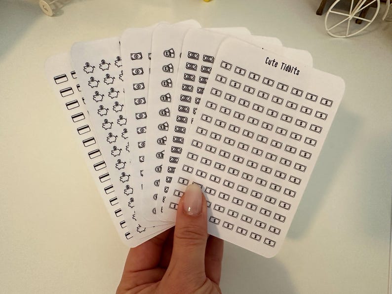 May include: A set of six sheets of stickers featuring various money-themed designs, including piggy banks, dollar bills, and stacks of money. The stickers are printed on white paper and have a black outline. The text "Cute Tidbits" is printed on one of the sheets.