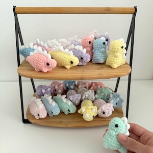 May include: A two-tiered display stand holds a collection of colorful, crocheted dinosaur toys. The dinosaurs are in various pastel colors, including pink, yellow, blue, and purple. One dinosaur is held in a hand. The stand has a bamboo handle and black metal frame.