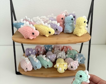 May include: A two-tiered display stand holds a collection of colorful, crocheted dinosaur toys. The dinosaurs are in various pastel colors, including pink, yellow, blue, and purple. One dinosaur is held in a hand. The stand has a bamboo handle and black metal frame.