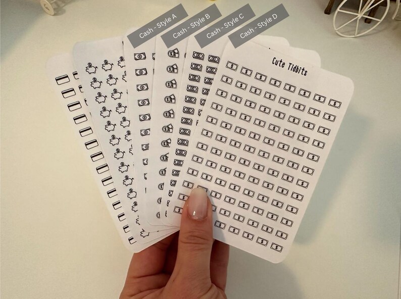 May include: A set of five sheets of black and white money themed stickers. The stickers are in four different styles, with the text "Cash - Style A", "Cash - Style B", "Cash - Style C", and "Cash - Style D" at the top of each sheet. The fifth sheet has the text "Cute Tidbits" at the top and features a grid of small money bag stickers.