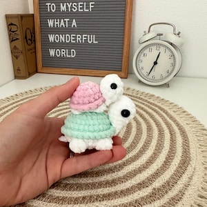 May include: A handmade crochet turtle in white, mint green, and pink, held in a hand. The turtle is on a beige and brown striped mat. In the background is a framed sign with the text "AND I THINK TO MYSELF WHAT A WONDERFUL WORLD" and a white alarm clock.