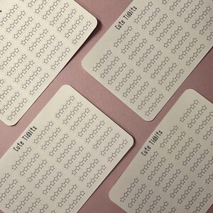 May include: Four white sticker sheets with rounded corners, each covered in rows of outlined star shapes. The text "Cute Tidbits" is printed in a handwritten style on each sheet. The stickers are arranged on a pink surface.