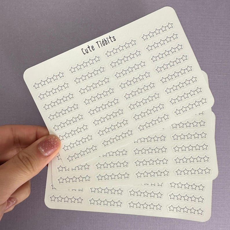 May include: A stack of white sticker sheets featuring rows of outlined star shapes. The text "Cute Tidbits" is printed in black at the top left. The stickers are held against a light purple background.