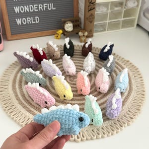 May include: A collection of small, crocheted dinosaur toys in various pastel colors, including blue, pink, yellow, and green, with white spines. The toys are arranged on a beige circular mat. The text "TO MYSELF WHAT A WONDERFUL WORLD" is visible in the background.