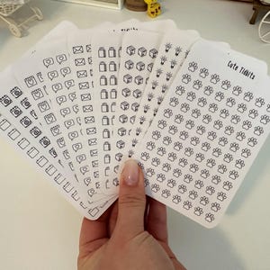 May include: A set of eight sheets of white sticker paper with black outline icons. The icons include calendars, cameras, hearts, mailboxes, shopping bags, boxes, light bulbs, and paw prints. The text "Cute Tidbits" is printed on one sheet.