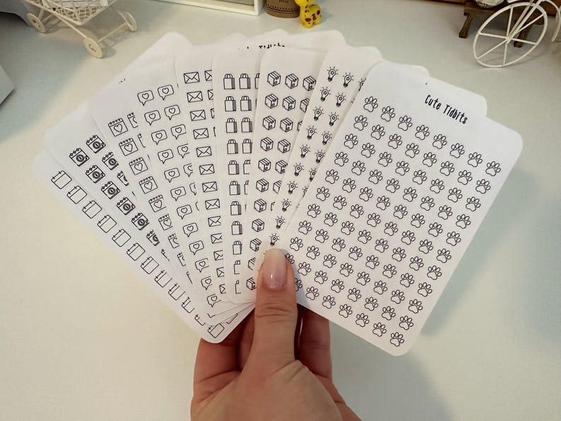 May include: A set of eight sheets of white sticker paper with black outline icons. The icons include calendars, cameras, hearts, mailboxes, shopping bags, boxes, light bulbs, and paw prints. The text "Cute Tidbits" is printed on one sheet.