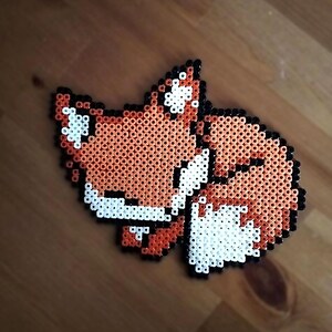 Op de afbeelding: Een knutselwerkje van strijkkralen in de vorm van een opgerolde vos. Het ontwerp bestaat uit oranje, witte en zwarte kralen die een slapende vos uitbeelden met de staart om het lichaam gewikkeld, omlijnd met zwarte kralen voor de vorm.