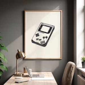 Könnte beinhalten: Schwarz-Weiß-Druck eines klassischen Nintendo Game Boy in einem Rahmen. Das Kunstwerk zeigt eine detaillierte Illustration der Handheld-Konsole mit ihren Knöpfen und dem Bildschirm. Der Druck hängt an einer grauen Wand über einem Schreibtisch.