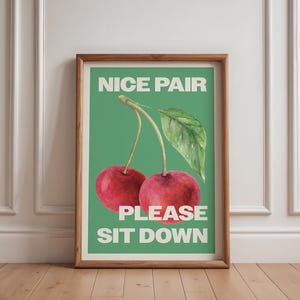 May include: A framed print with a green background and a watercolor illustration of two red cherries with a green stem and leaf. The text "NICE PAIR PLEASE SIT DOWN" is printed in white on the print.