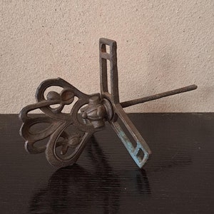 Puede incluir: Un soporte para plantas de metal vintage con un diseño decorativo y ornamentado. El soporte tiene un solo tallo central y una base con un diseño en forma de cruz.