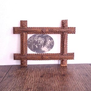Puede incluir: Un marco de fotos rectangular de madera vintage con una fotografía ovalada en blanco y negro. El marco está hecho de pequeñas piezas de madera muy juntas, creando una apariencia texturizada y rústica. Se encuentra sobre una superficie de madera oscura.
