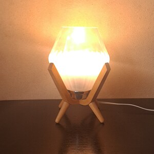 Peut inclure: Un abat-jour de lampe en verre blanc avec un motif géométrique repose sur un pied en bois à quatre pieds. La lampe est allumée et émet une lumière chaude et jaune.