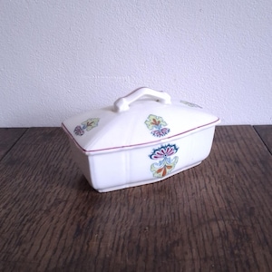 May include: A white rectangular ceramic butter dish with a lid and handle. The dish is decorated with colorful floral patterns and a red trim. The butter dish is sitting on a dark wooden surface.