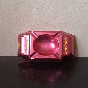 Può includere: Un posacenere in metallo rosa con la scritta "AIR-INDIA" in lettere gialle sul lato.